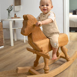 A little toddler sat on a wooden rocking horse, wearing the Lassig organic cotton muslin jumpsuit in a natural cream colour.