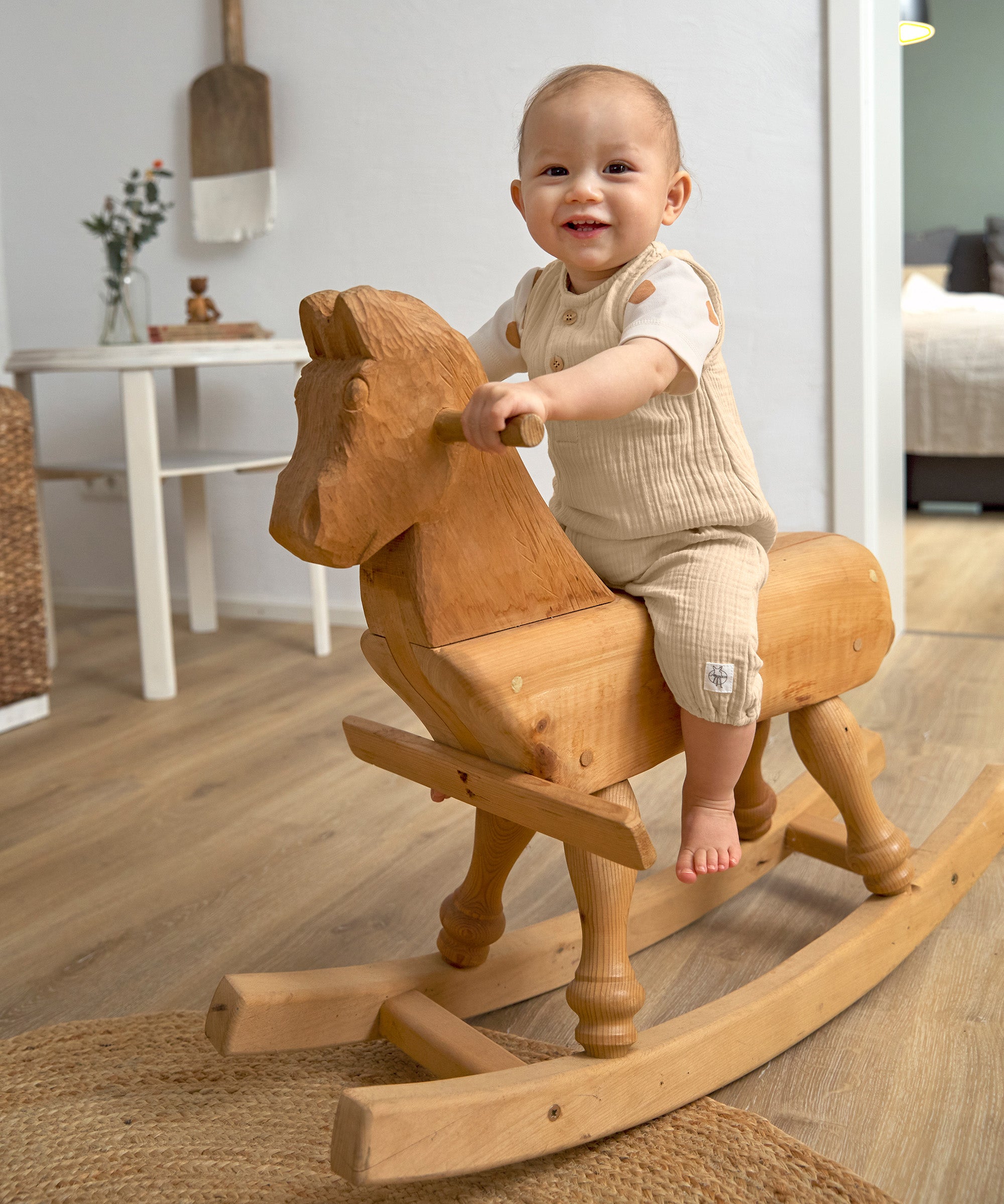 A little toddler sat on a wooden rocking horse, wearing the Lassig organic cotton muslin jumpsuit in a natural cream colour.