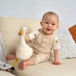 A baby sat on a sofa, holding an organic stuffed toy and wearing the Lassig organic cotton muslin jumpsuit in a natural cream colour.