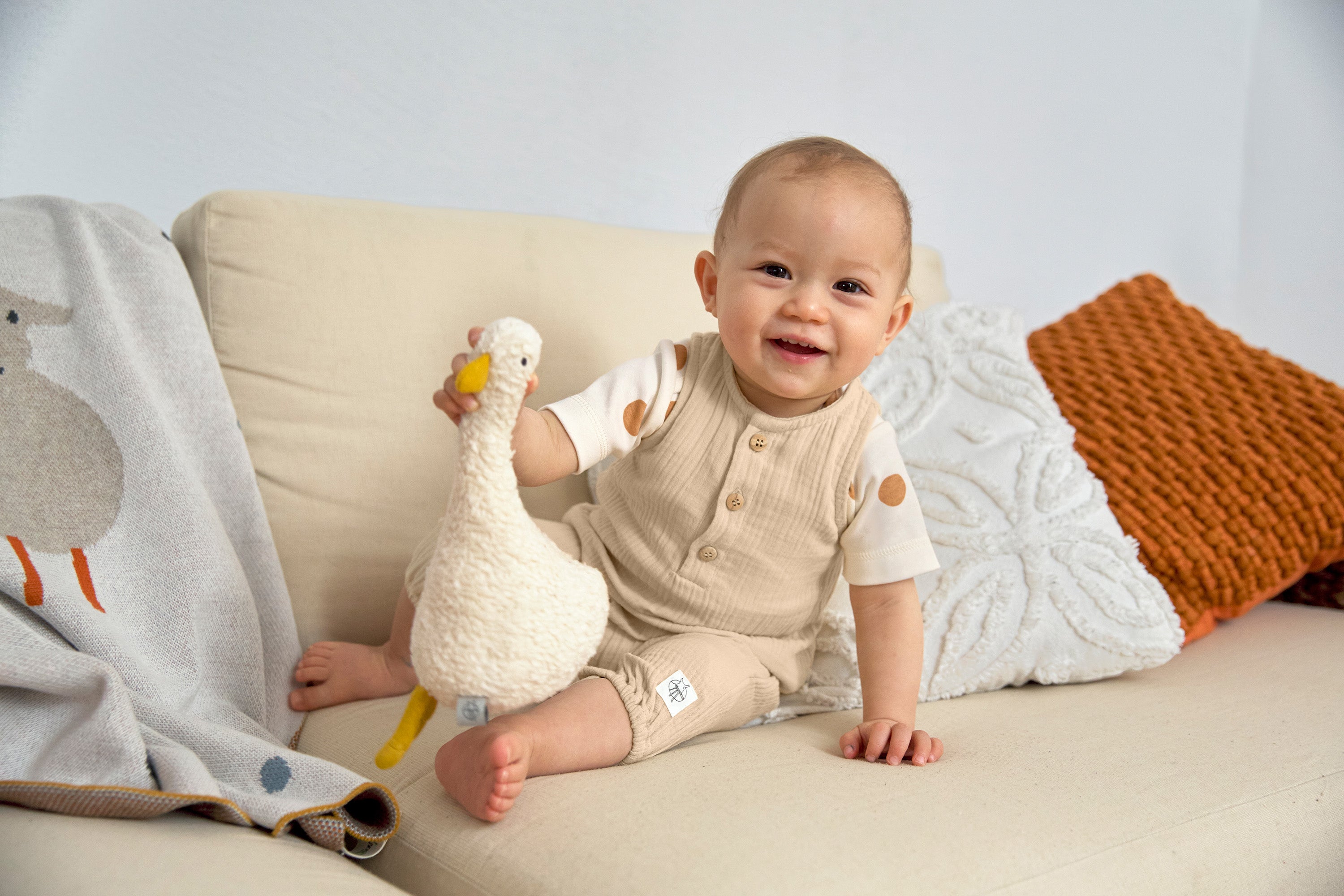 A baby sat on a sofa, holding an organic stuffed toy and wearing the Lassig organic cotton muslin jumpsuit in a natural cream colour.