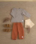 An organic cotton muslin outfit from Lassig, laid out on the floor. It includes the Lassig muslin pants in orange rust. 