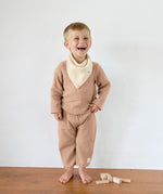 A boy in an organic cotton muslin outfit from Lassig, with the Bandana Dribble Bib in a cream Vanilla colour. 