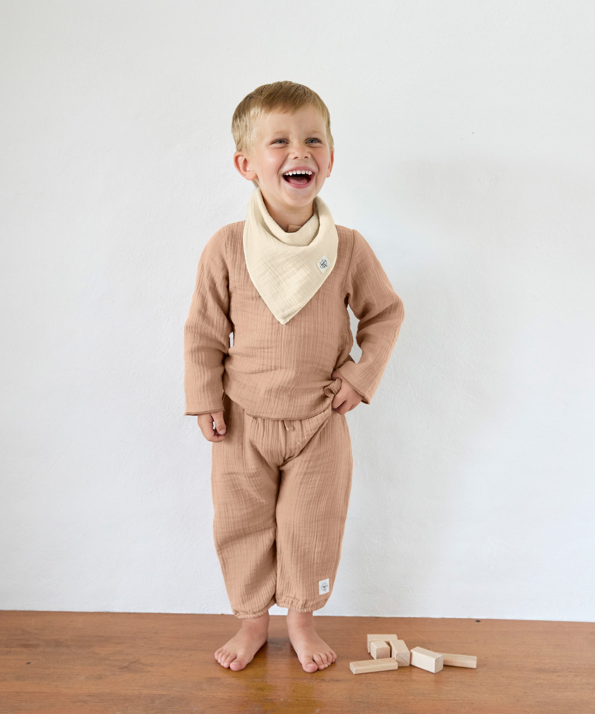 A boy in an organic cotton muslin outfit from Lassig, with the Bandana Dribble Bib in a cream Vanilla colour. 