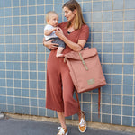 A woman carrying her baby and holding the Lassig rolltop nappy change backpack in cinnamon
