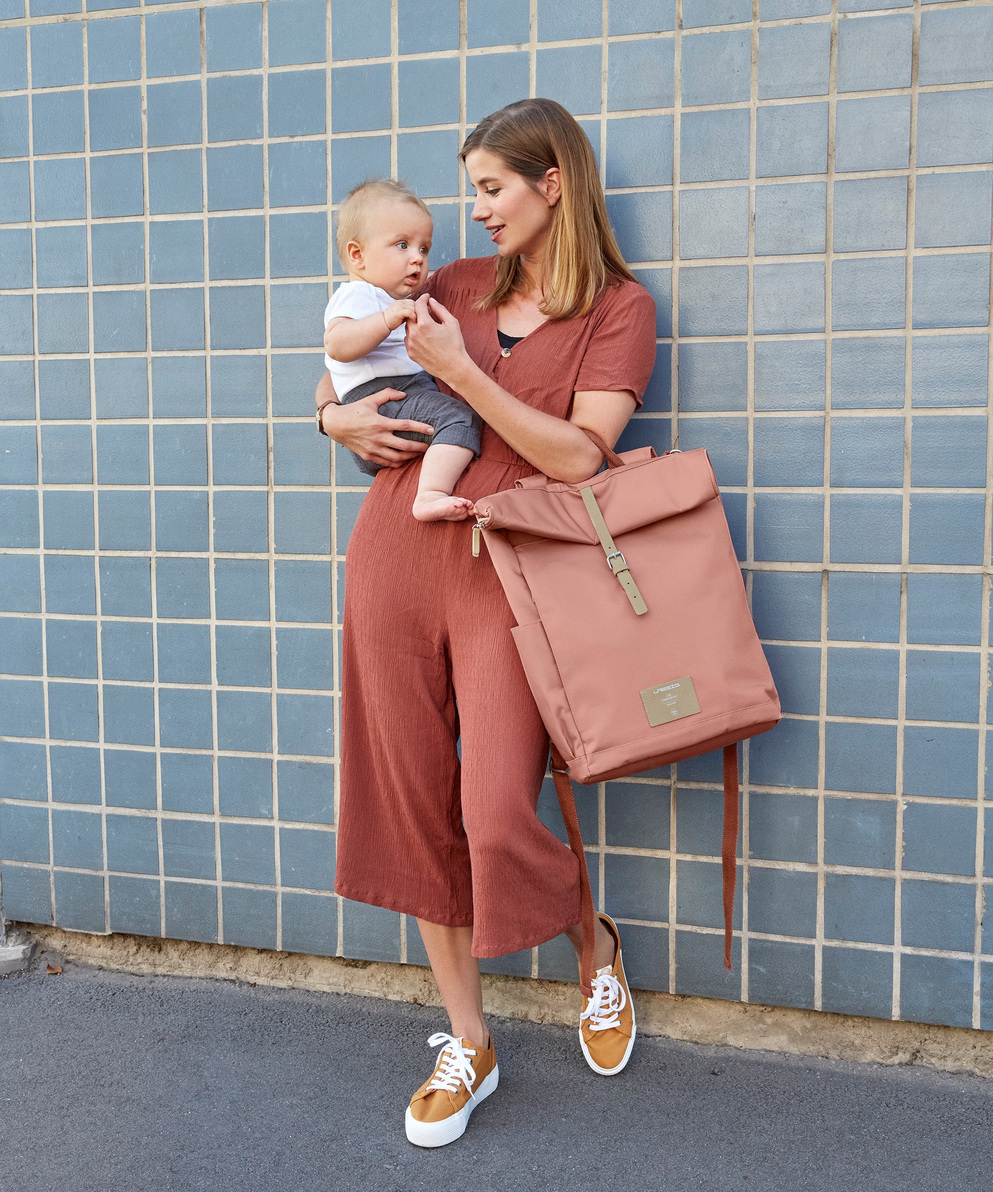 A woman carrying her baby and holding the Lassig rolltop nappy change backpack in cinnamon