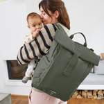 A woman holding her baby, and carrying the Lassig Rolltop Up nappy change backpack in olive green.