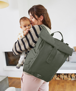 A woman holding her baby, and carrying the Lassig Rolltop Up nappy change backpack in olive green.