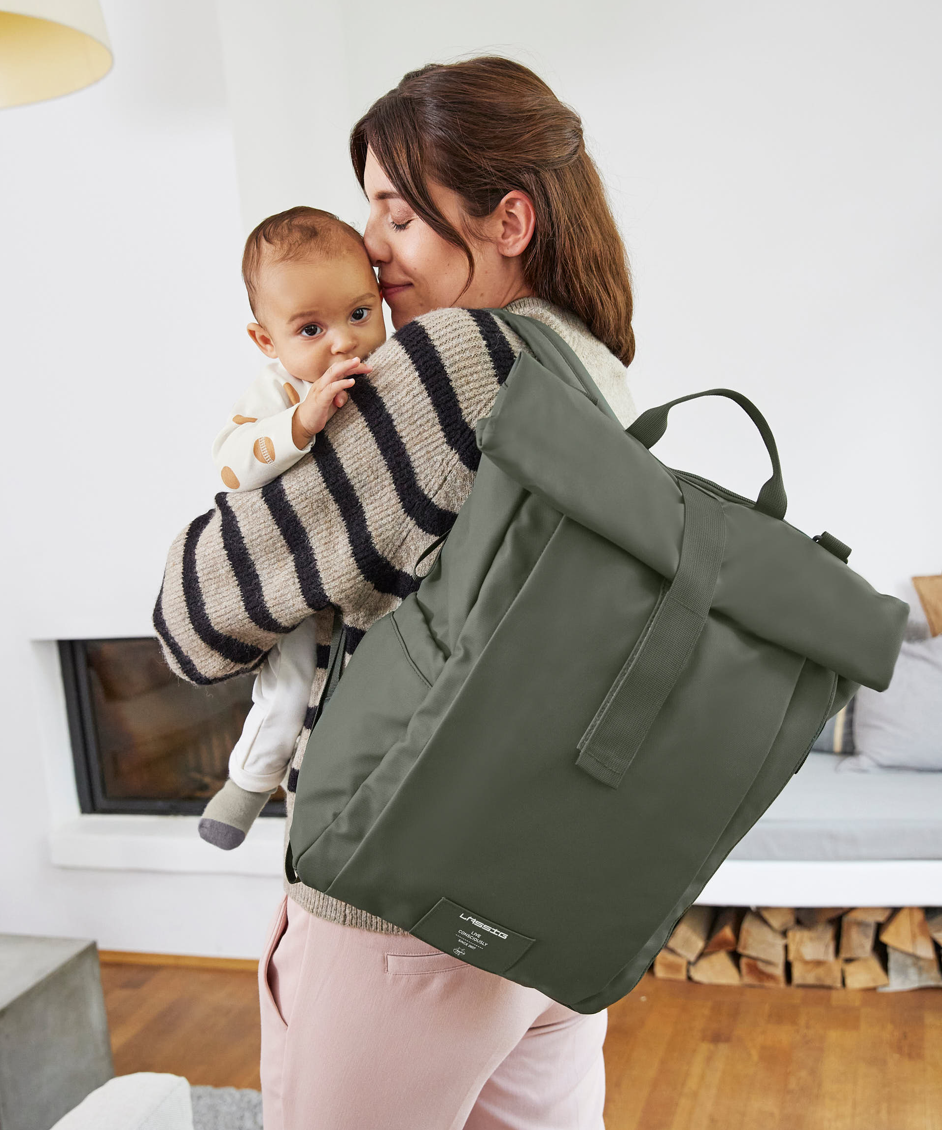 A woman holding her baby, and carrying the Lassig Rolltop Up nappy change backpack in olive green.