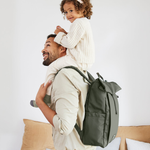 A man carrying his child on his shoulders, and wearing the Lassig Rolltop Up nappy change backpack in olive green.
