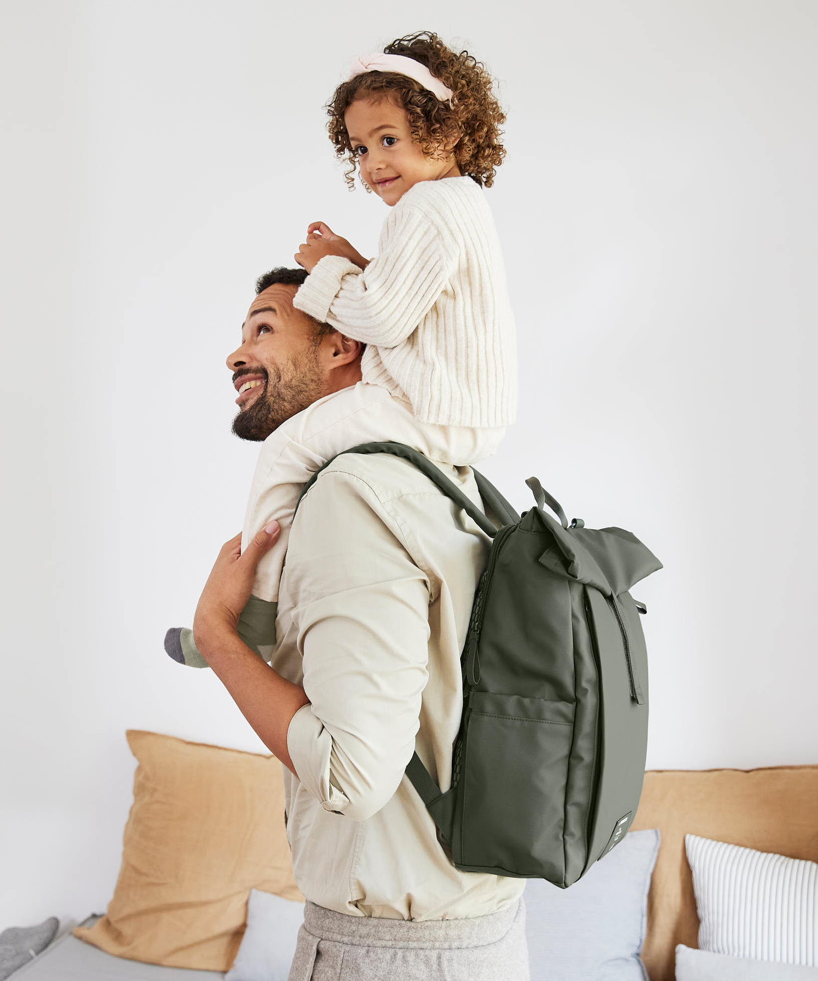 A man carrying his child on his shoulders, and wearing the Lassig Rolltop Up nappy change backpack in olive green.