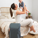 A woman holding up her baby, with the Lassig Slender Up nappy change backpack in reflective anthracite on the floor next to them.