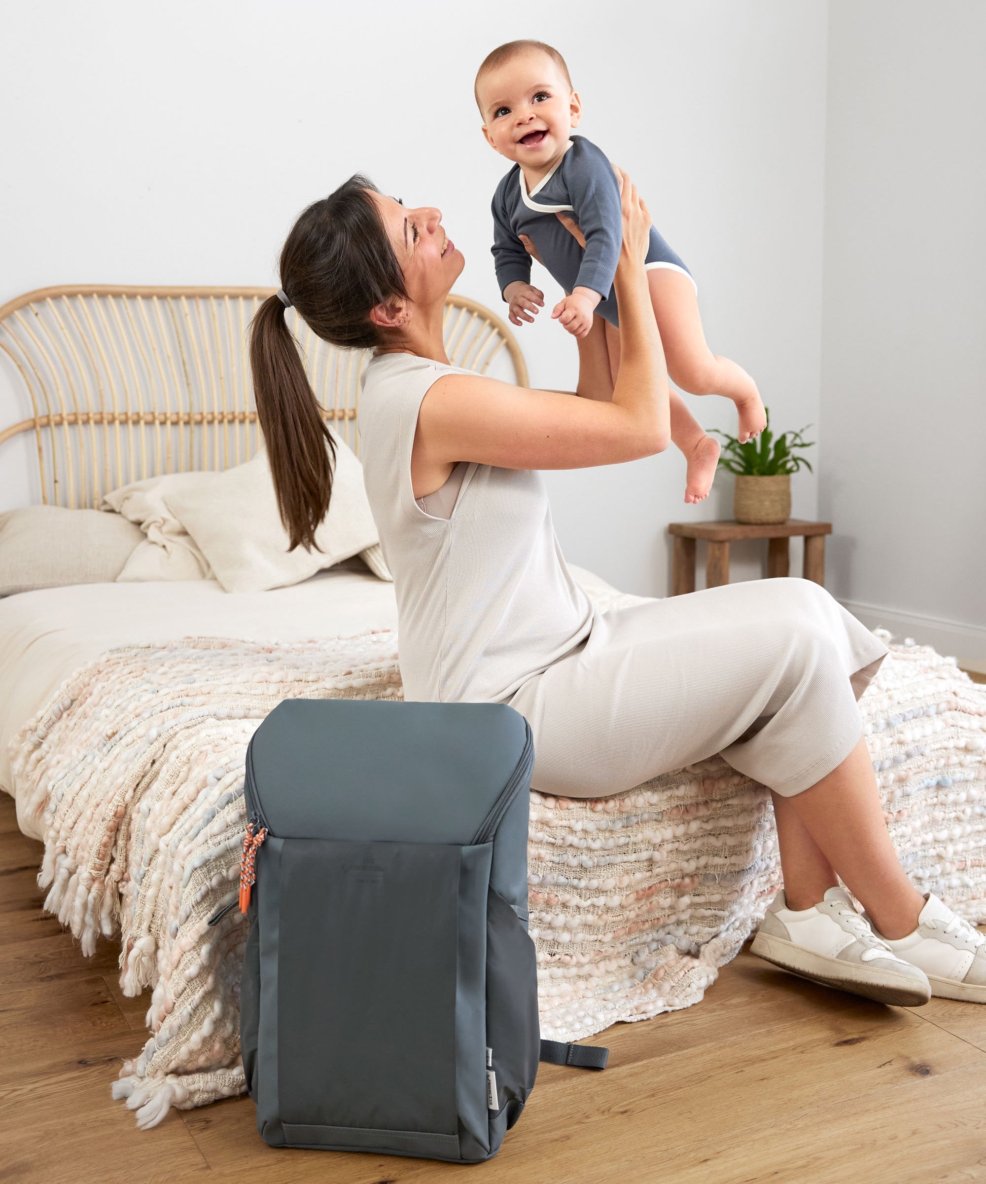 A woman holding up her baby, with the Lassig Slender Up nappy change backpack in reflective anthracite on the floor next to them.