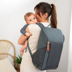 A woman holding her baby, and wearing the Lassig Slender Up nappy change backpack in reflective anthracite.