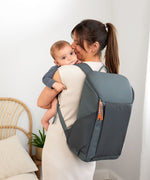 A woman holding her baby, and wearing the Lassig Slender Up nappy change backpack in reflective anthracite.