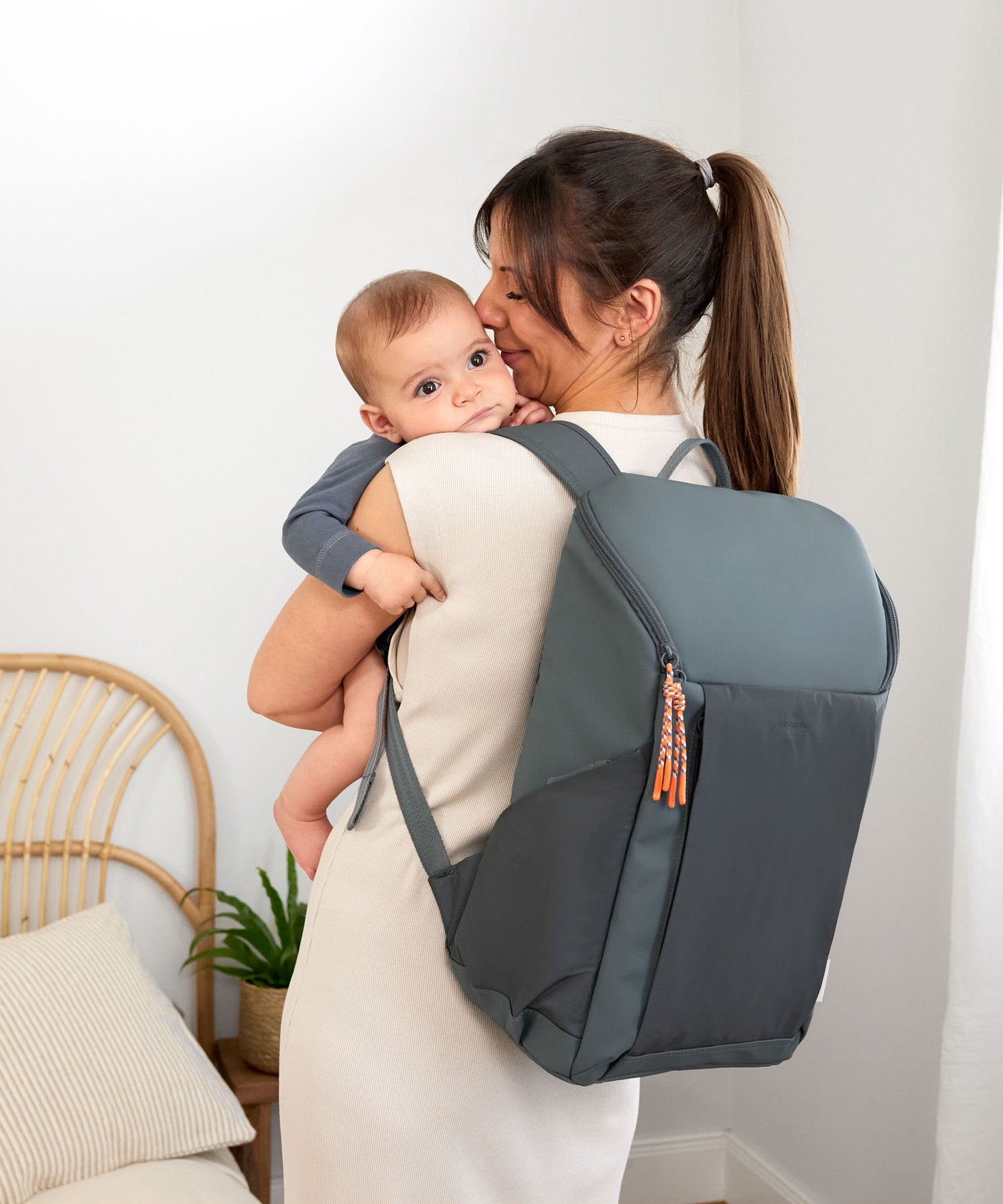A woman holding her baby, and wearing the Lassig Slender Up nappy change backpack in reflective anthracite.