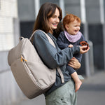 A mother carrying her baby, and wearing the Lassig Slender Up nappy change backpack in Taupe.
