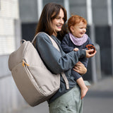 A mother carrying her baby, and wearing the Lassig Slender Up nappy change backpack in Taupe.