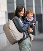 A mother carrying her baby, and wearing the Lassig Slender Up nappy change backpack in Taupe.