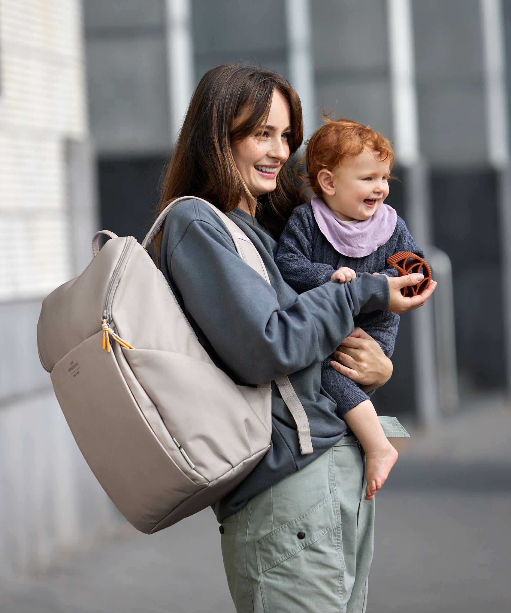 A mother carrying her baby, and wearing the Lassig Slender Up nappy change backpack in Taupe.