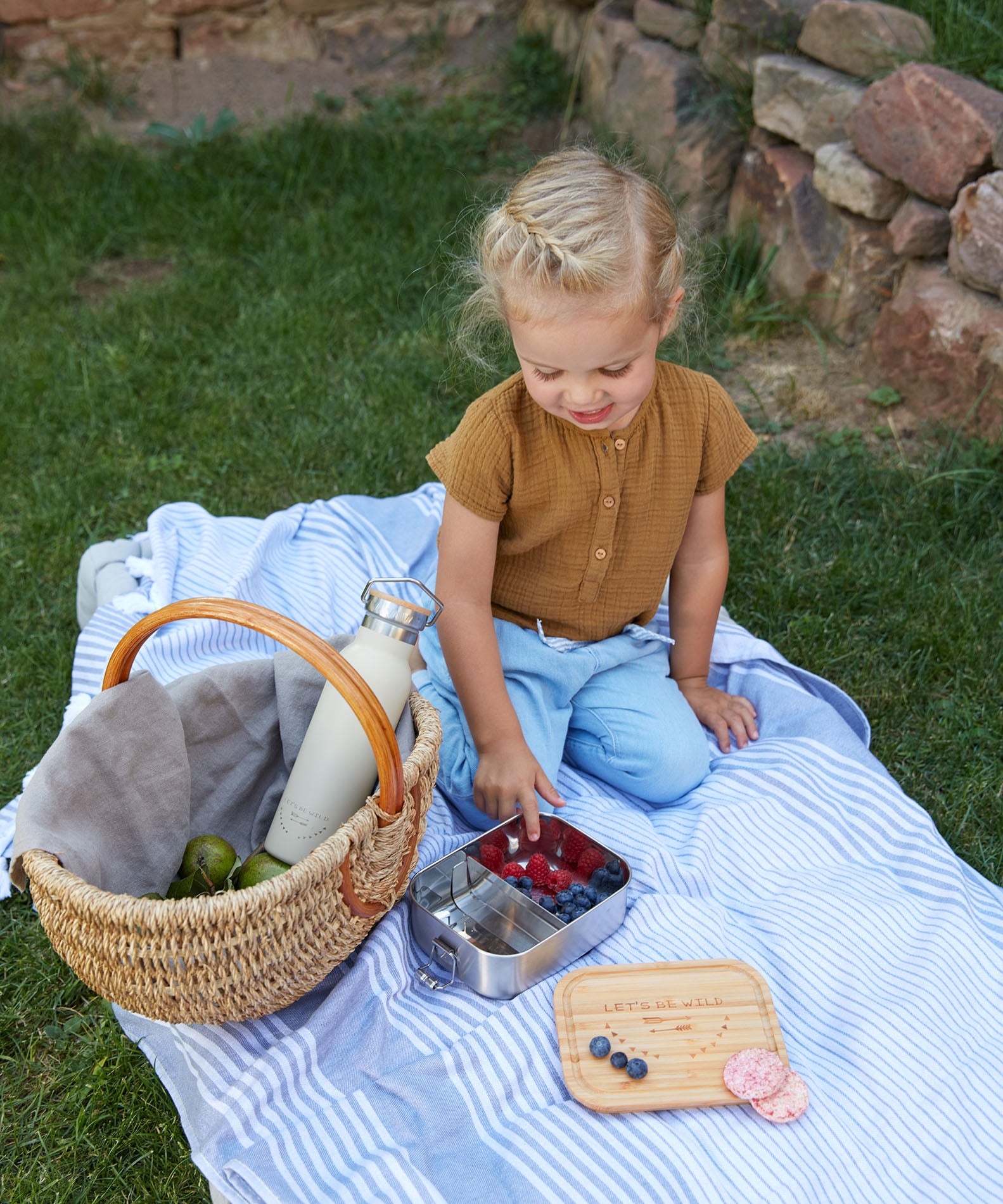 A child sitting outdoors eating berries from a Lassig Stainless Steel Bamboo Lunchbox. This stainless steel lunchbox is part of a wide range of reusable food containers available at Babipur. 
