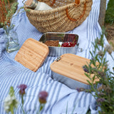The Lassig Stainless Steel Bamboo Lunchbox with a Garden Explorer design placed on a striped blanket outdoors. Another design can be seen in the background. This stainless steel lunchbox is part of a wide range of reusable food containers available at Babipur. 