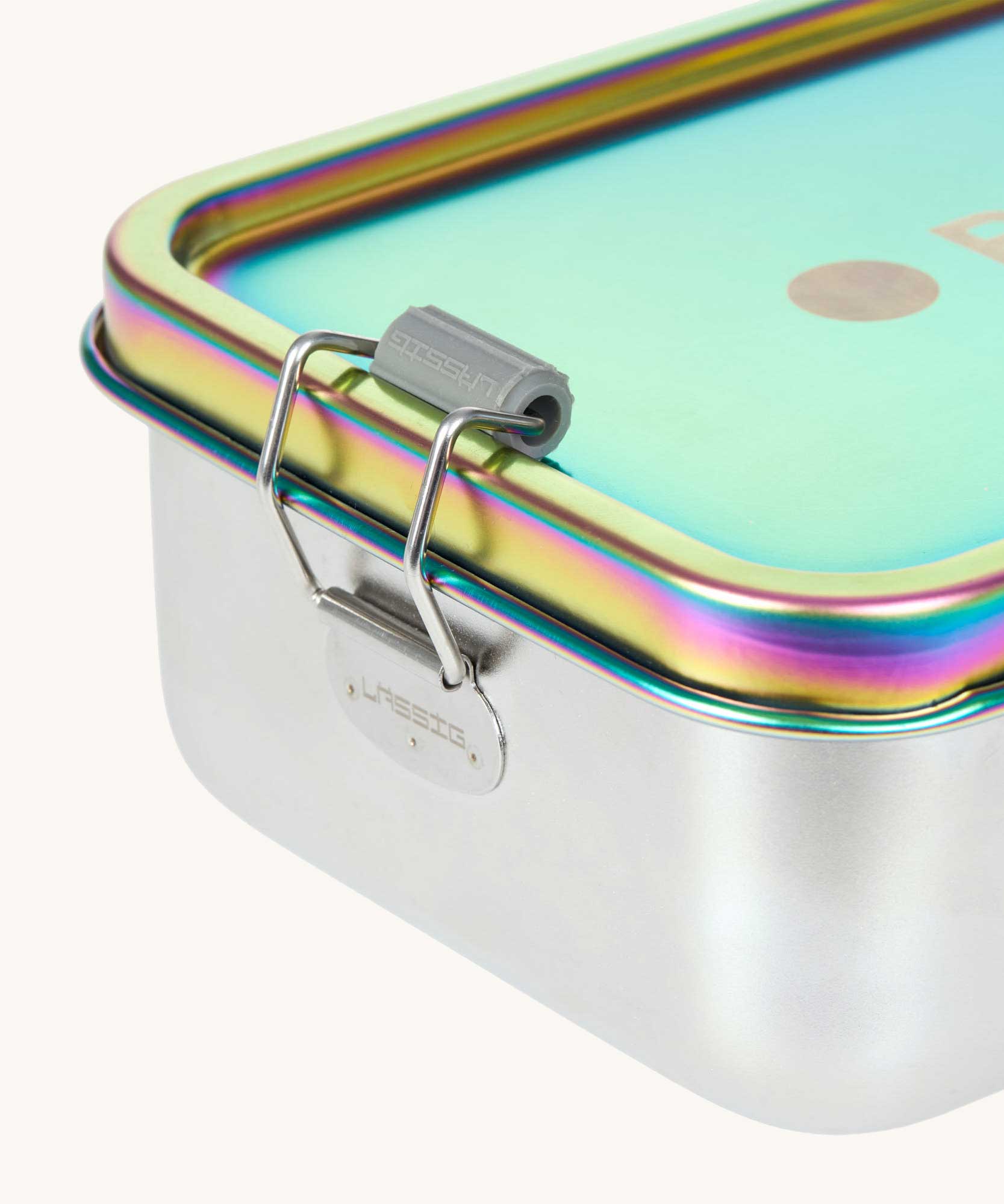 A close up of the clamp latch closure on the Lassig Stainless Steel Lunchbox in a  Solid Bold Rainbow design. A stainless steel lunchbox with removable divider featuring an iridescent rainbow coloured lid. This lunchbox is part of a wide range of reusable items available at Babipur. 