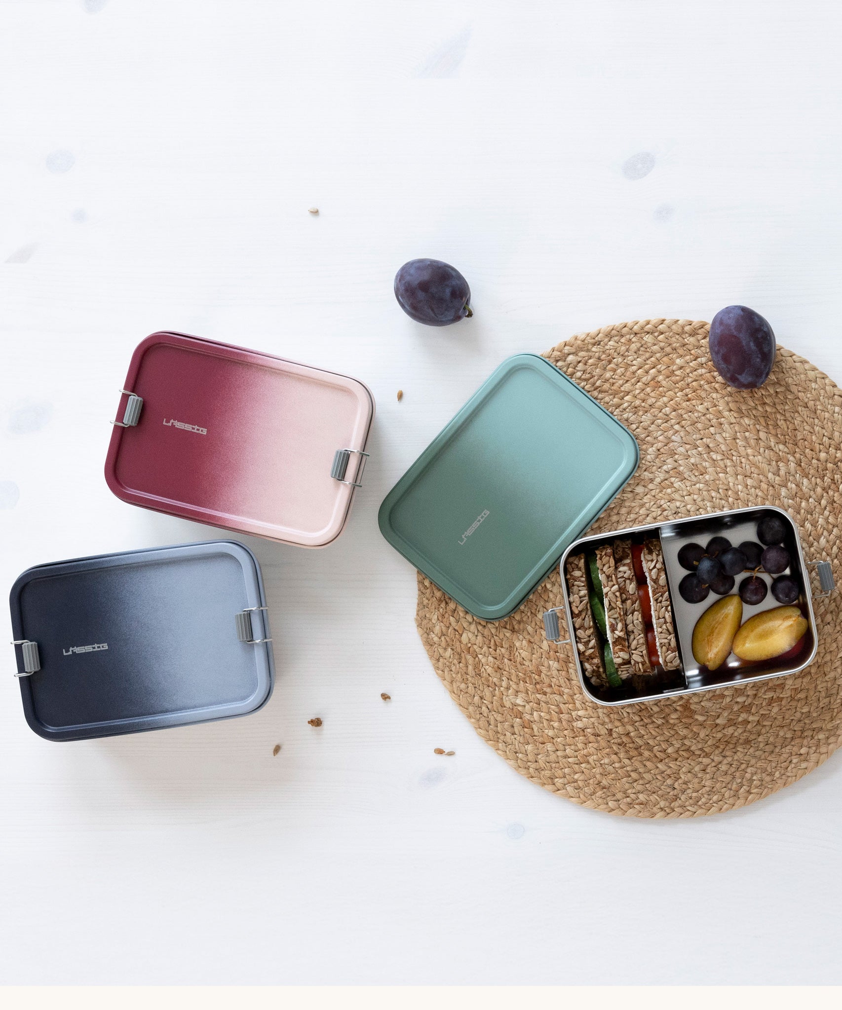 A collection of three Lassig Stainless Steel Lunchbox. A Unique Olive Green colour shown with the lid off and filled with food and a berry rose pink and a dark blue colour box with lids on. 