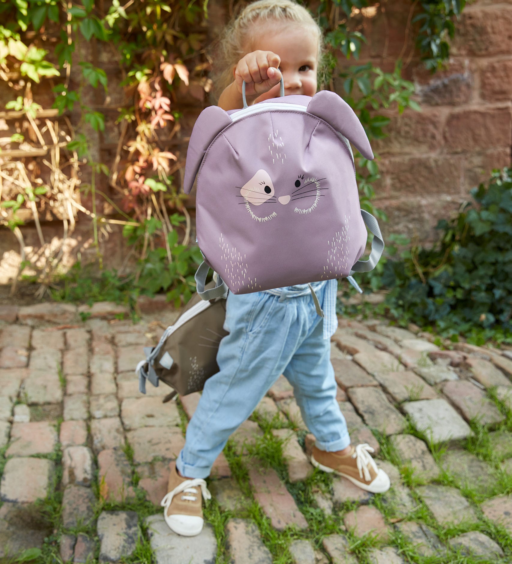 A child standing on a cobbled path and holding the Lassig bunny tiny backpack by the loop handle on the top