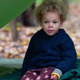 A child wearing the Little Green Radicals ginger navy knitted jumper in woodland under a shelter. A thick cable knit style jumper in dark navy GOTS organic cotton fabric with thick striped cuffs.