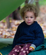 A child wearing the Little Green Radicals ginger navy knitted jumper in woodland under a shelter. A thick cable knit style jumper in dark navy GOTS organic cotton fabric with thick striped cuffs.