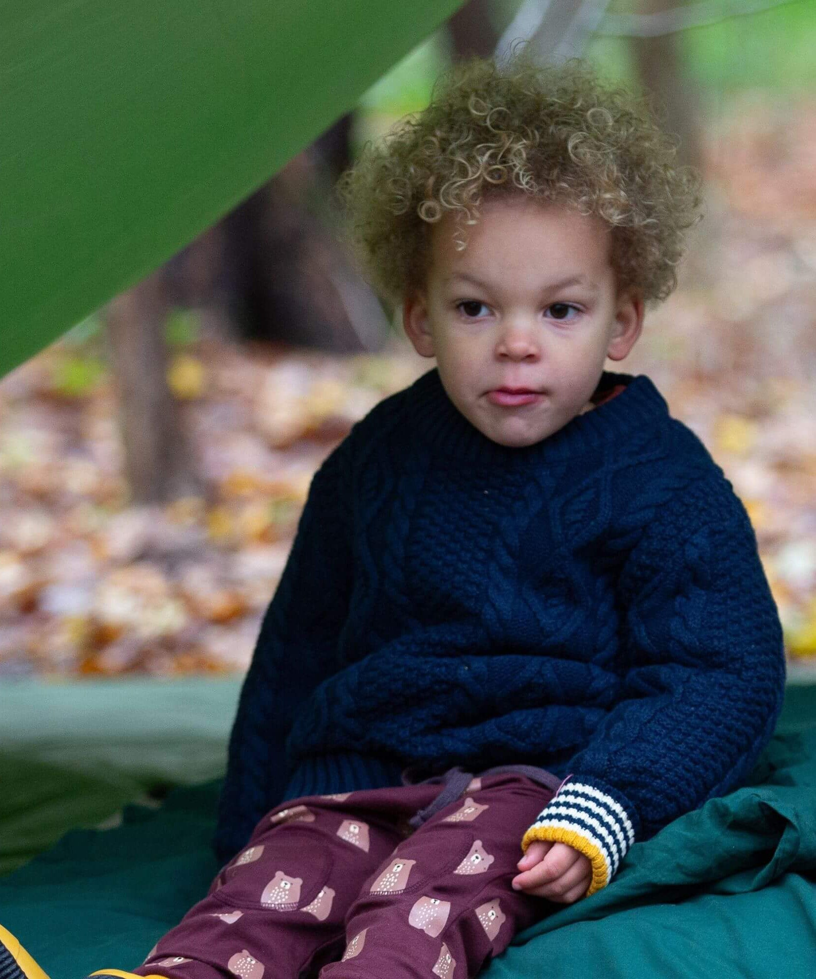A child wearing the Little Green Radicals ginger navy knitted jumper in woodland under a shelter. A thick cable knit style jumper in dark navy GOTS organic cotton fabric with thick striped cuffs.