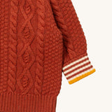 A close up of the Little Green Radicals ginger aran knitted jumper available at Babipur. Showing the cable knit and the striped cuff detail on dark orange organic cotton fabric