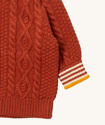 A close up of the Little Green Radicals ginger aran knitted jumper available at Babipur. Showing the cable knit and the striped cuff detail on dark orange organic cotton fabric
