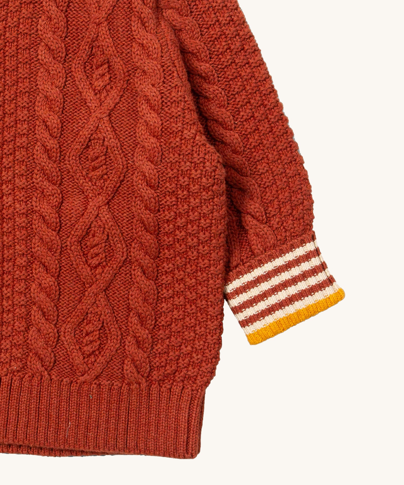 A close up of the Little Green Radicals ginger aran knitted jumper available at Babipur. Showing the cable knit and the striped cuff detail on dark orange organic cotton fabric
