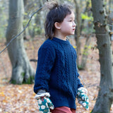 A child standing in woodland wearing the Little Green Radicals navy aran knitted jumpe. A thick cable knit style jumper in dark navy GOTS organic cotton fabric with striped cuffs.