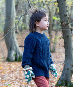 A child standing in woodland wearing the Little Green Radicals navy aran knitted jumpe. A thick cable knit style jumper in dark navy GOTS organic cotton fabric with striped cuffs.