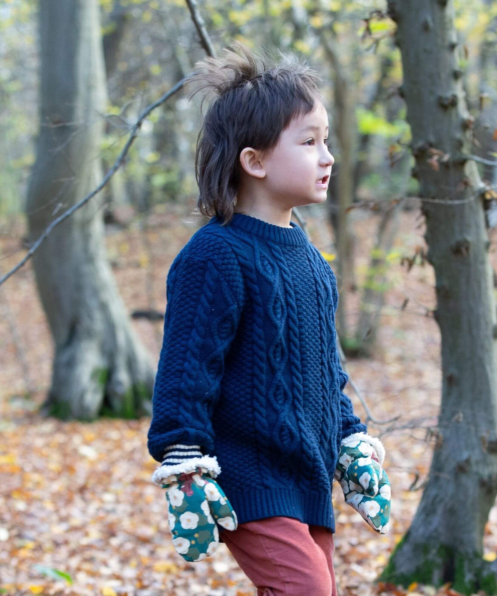 A child standing in woodland wearing the Little Green Radicals navy aran knitted jumpe. A thick cable knit style jumper in dark navy GOTS organic cotton fabric with striped cuffs.