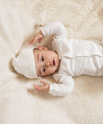 A baby lying down on a soft surface wearing the Little Green Radicals 100% organic cotton footed babygrow and the baby hat
