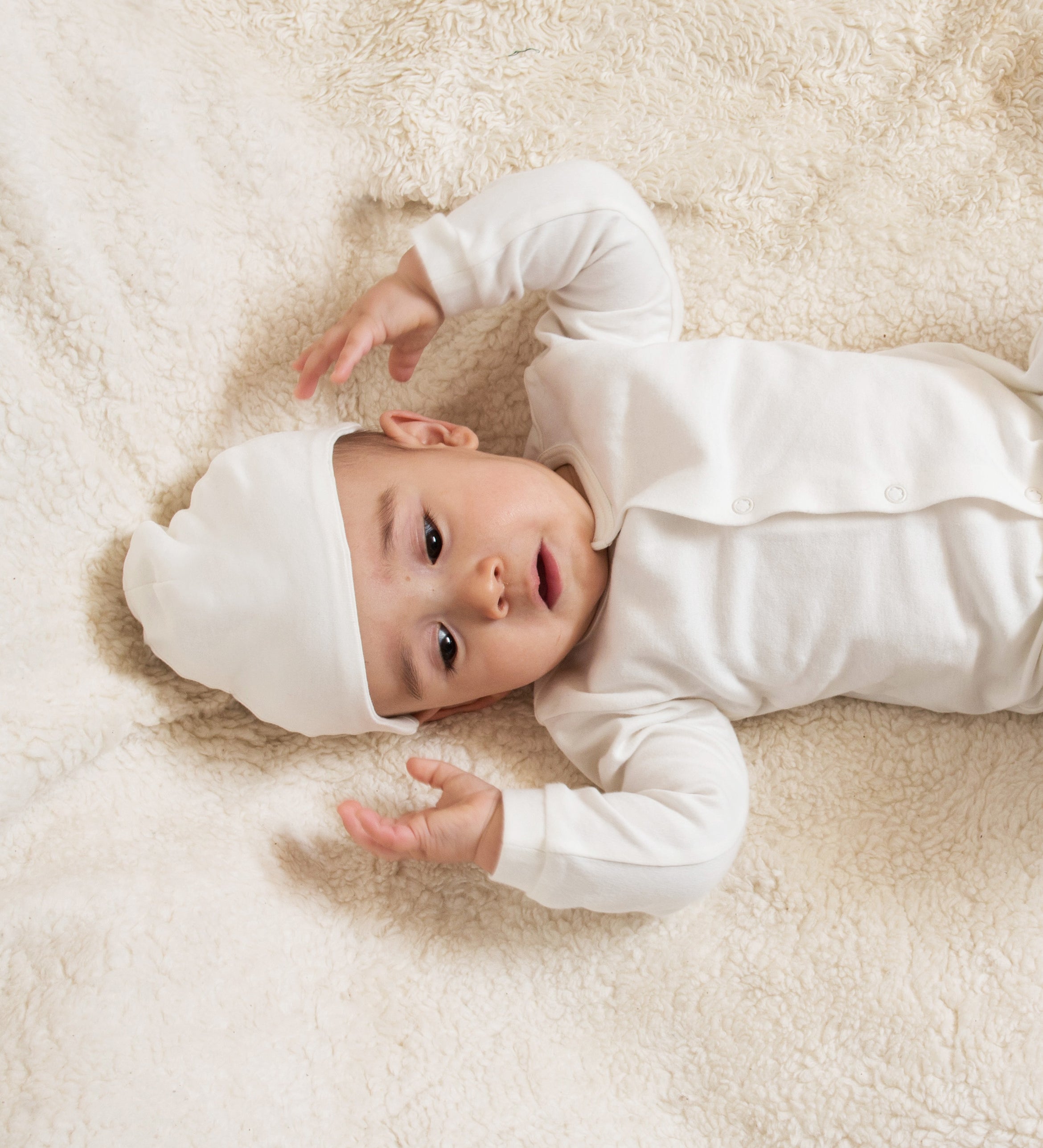 A baby lying down on a soft surface wearing the Little Green Radicals 100% organic cotton footed babygrow and the baby hat