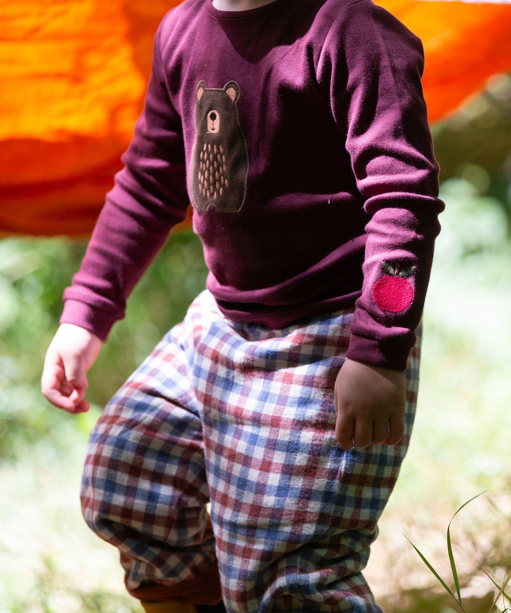 A close up of a child standing outside and wearing the dark maroon Little Green Radicals teddy bear applique long sleeve t shirt showing the apple applique detail. 