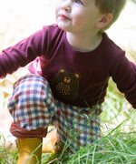 A close up of a child playing in grass and wearing the dark maroon Little Green Radicals teddy bear applique long sleeve t shirt showing the brown bear applique detail.