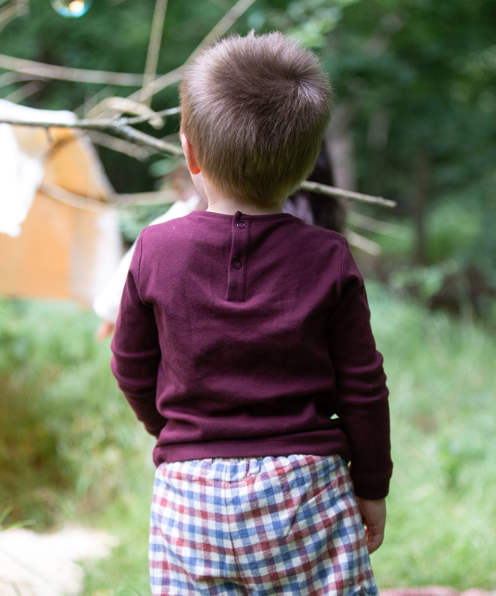 A child standing outside and showing the back of the dark maroon Little Green Radicals teddy bear applique long sleeve t shirt showing the popper fastener detail