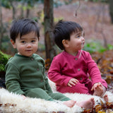 2 children sitting outside on a fluffy rug on leaves and wearing the Little Green Radicals holly berry pink and green chive waffle babygrows available at Babipur. A long sleeved, footless all in one suit made from GOTS organic cotton fabric with button fasteners and a small pouch chest pocket