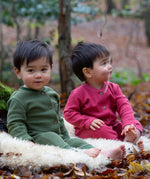 2 children sitting outside on a fluffy rug on leaves and wearing the Little Green Radicals holly berry pink and green chive waffle babygrows available at Babipur. A long sleeved, footless all in one suit made from GOTS organic cotton fabric with button fasteners and a small pouch chest pocket