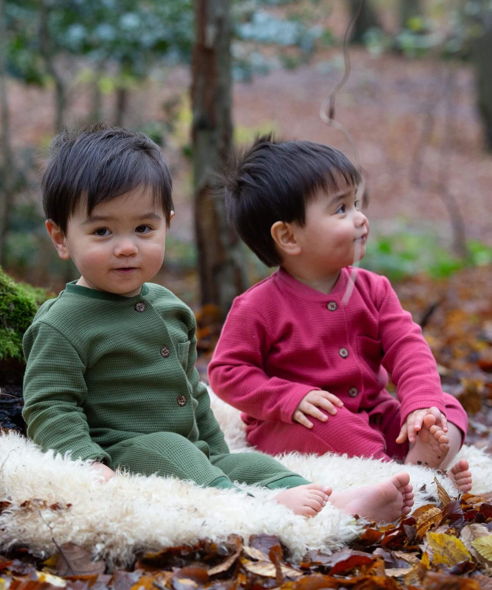2 children sitting outside on a fluffy rug on leaves and wearing the Little Green Radicals holly berry pink and green chive waffle babygrows available at Babipur. A long sleeved, footless all in one suit made from GOTS organic cotton fabric with button fasteners and a small pouch chest pocket