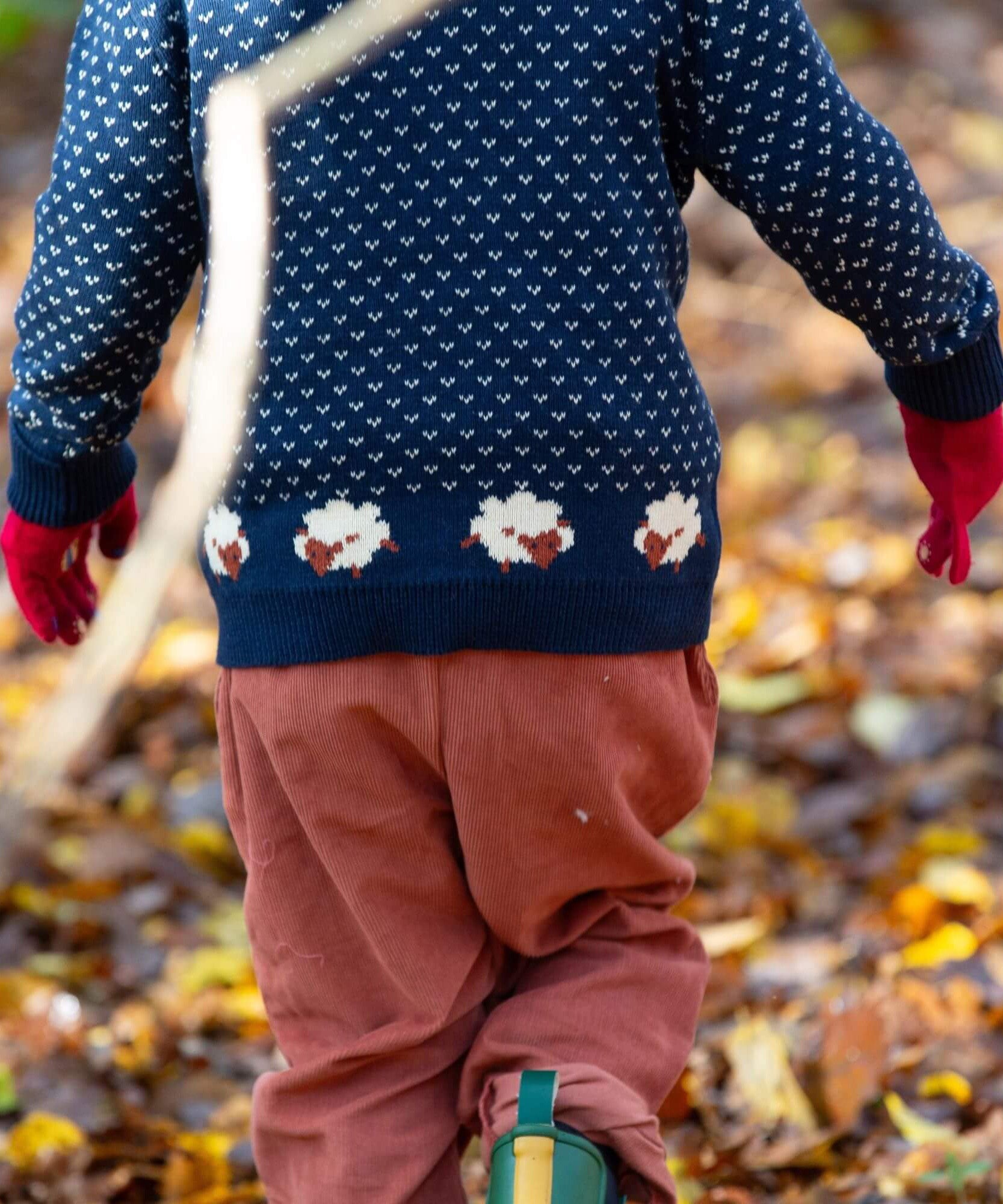 A child walking through leaves in a woodland  and wearing the Little Green Radicals comfy corduroy trousers in ginger with the navy sheep knitted jumper.  A corduroy loose fitted trousers in dark orange organic cotton fabric