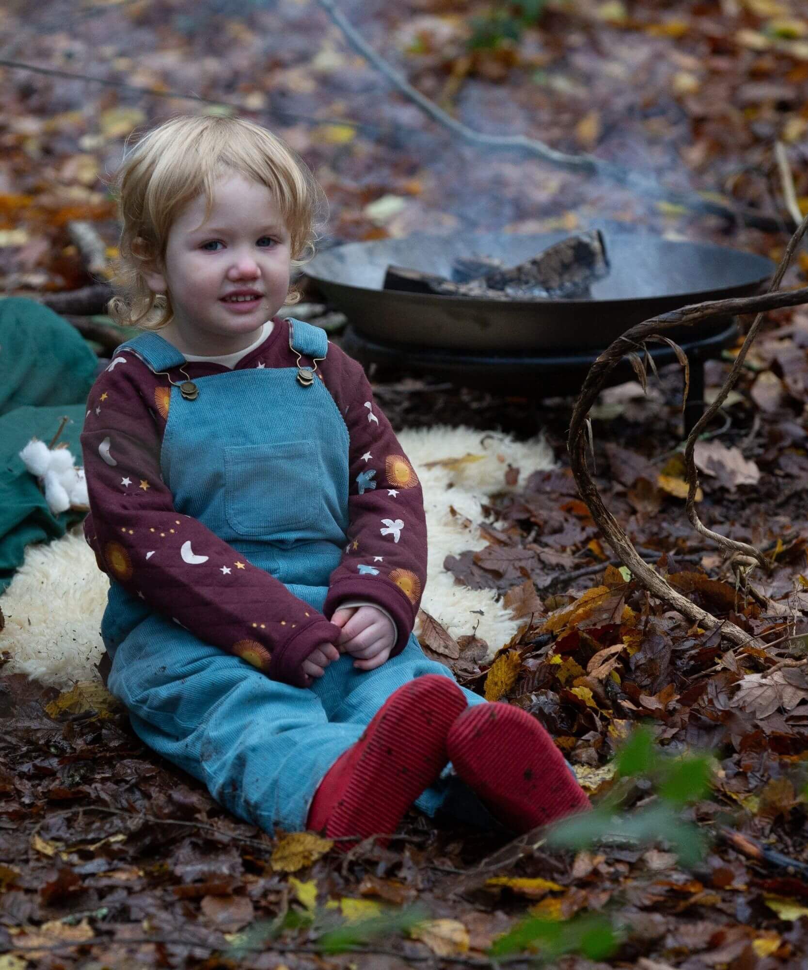 A child sitting down surrounded by leaves and wearing the Little Green Radicals pale blue lined corduroy dungarees