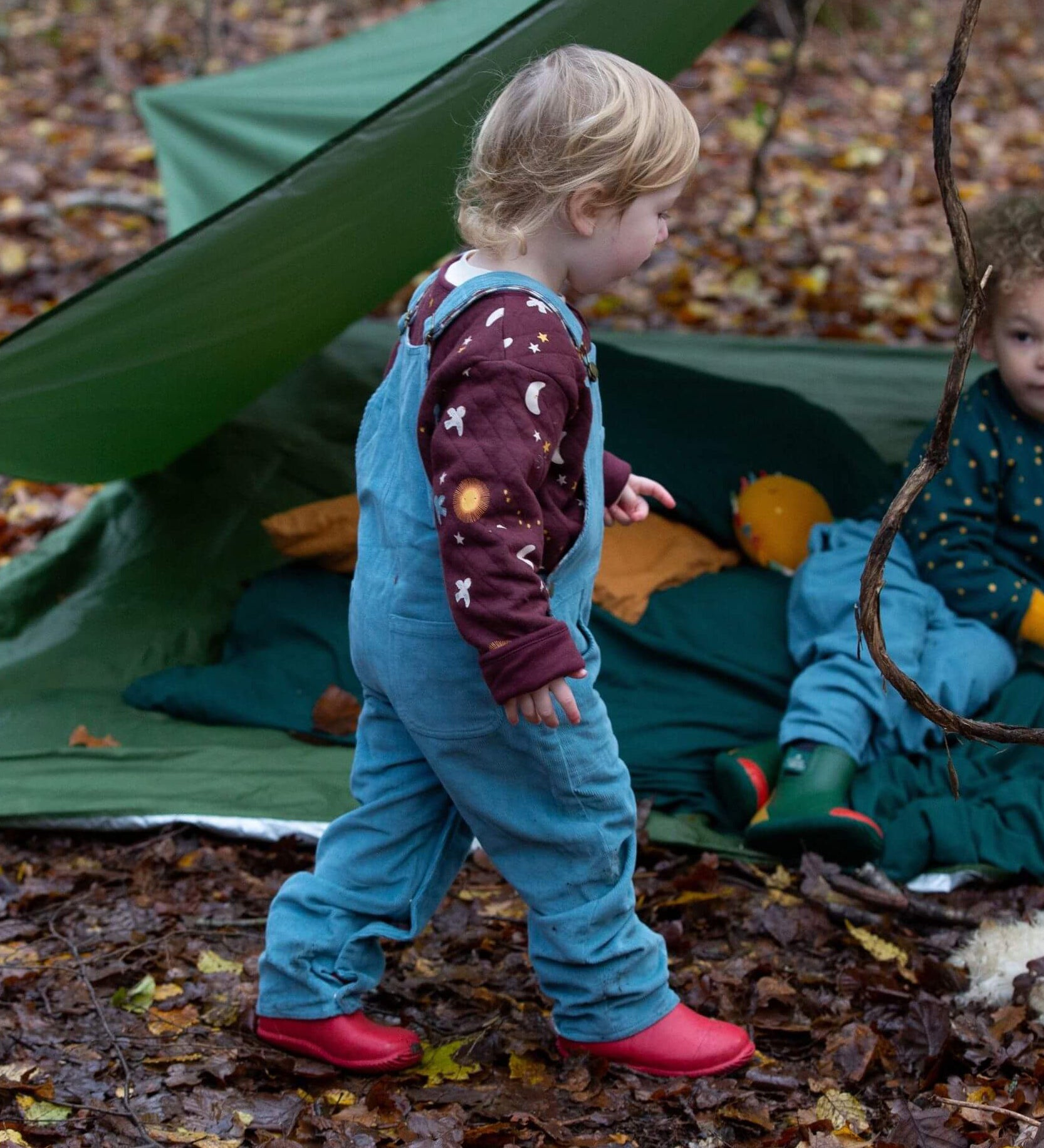 A child walking in the woods and wearing the Little Green Radicals pale blue lined corduory dungarees over the quilted flying birds sweater.
