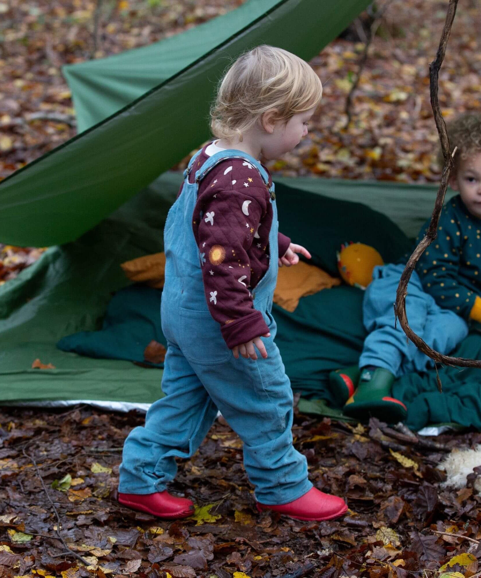 A child walking in the woods and wearing the Little Green Radicals pale blue lined corduory dungarees over the quilted flying birds sweater.