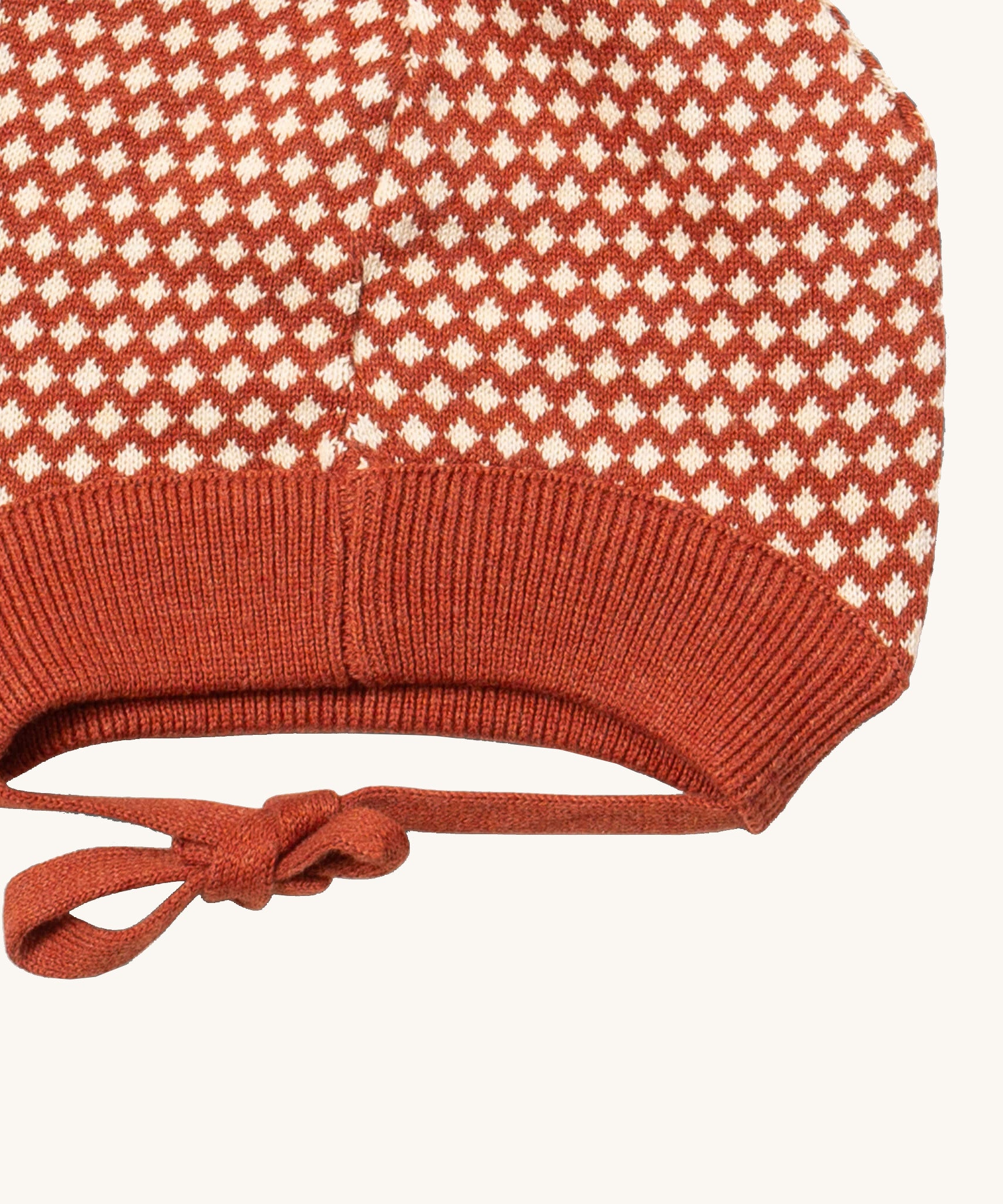 A close up of the Little Green Radicals ginger diamond knitted beanie hat showing the closed dark orange tie detail on organic cotton fabric